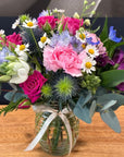A vibrant mixed floral arrangement in a clear glass jar with a white ribbon, displayed on a wooden tabletop. The bouquet features pink carnations, pink and magenta roses, white daisies, purple flowers, blue thistles, and green foliage.