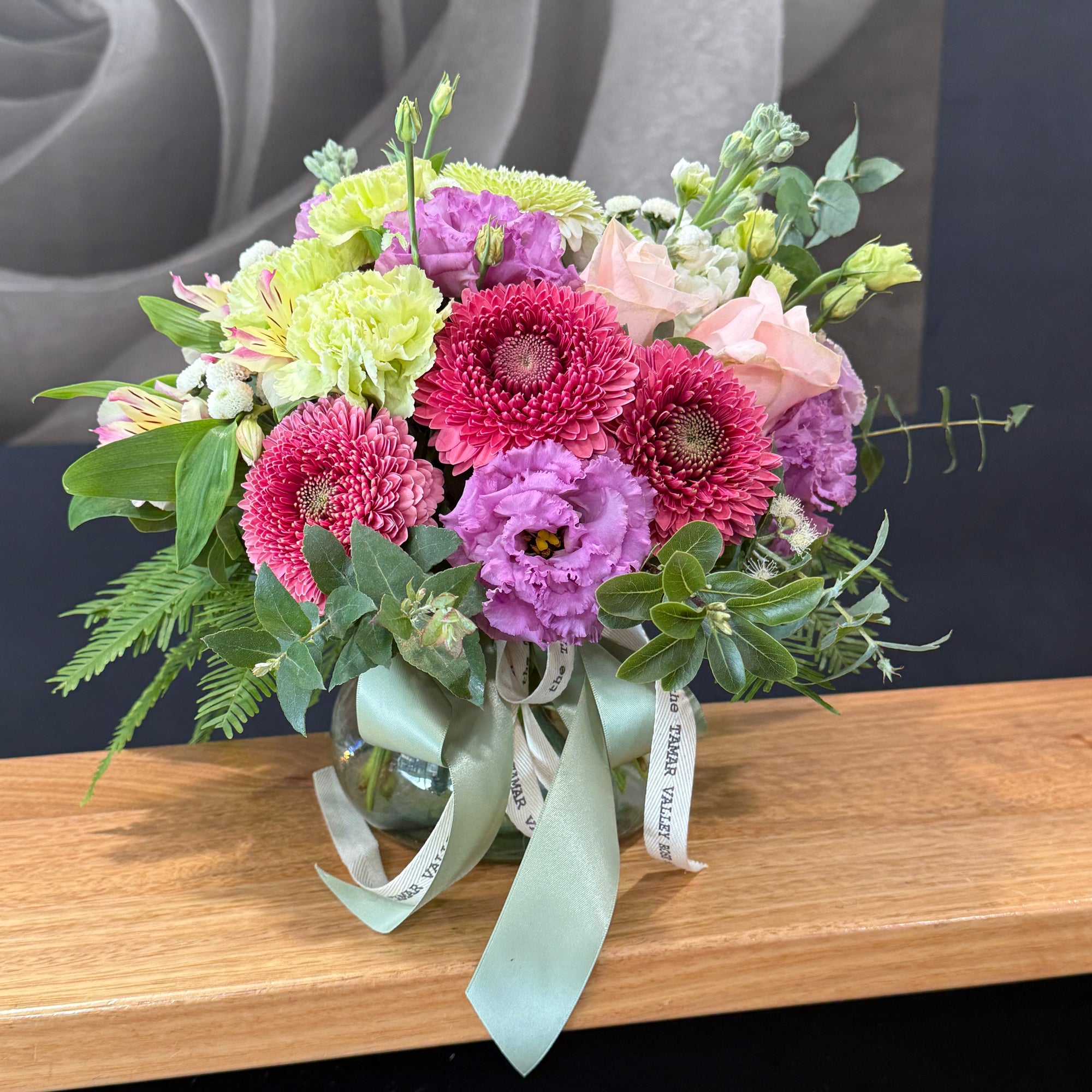 A vibrant floral arrangement in a clear glass vase, featuring pink and purple flowers, light green blooms, and lush green foliage, tied with a pale green satin ribbon and a white paper band.