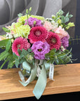A vibrant floral arrangement in a clear glass vase, featuring pink and purple flowers, light green blooms, and lush green foliage, tied with a pale green satin ribbon and a white paper band.