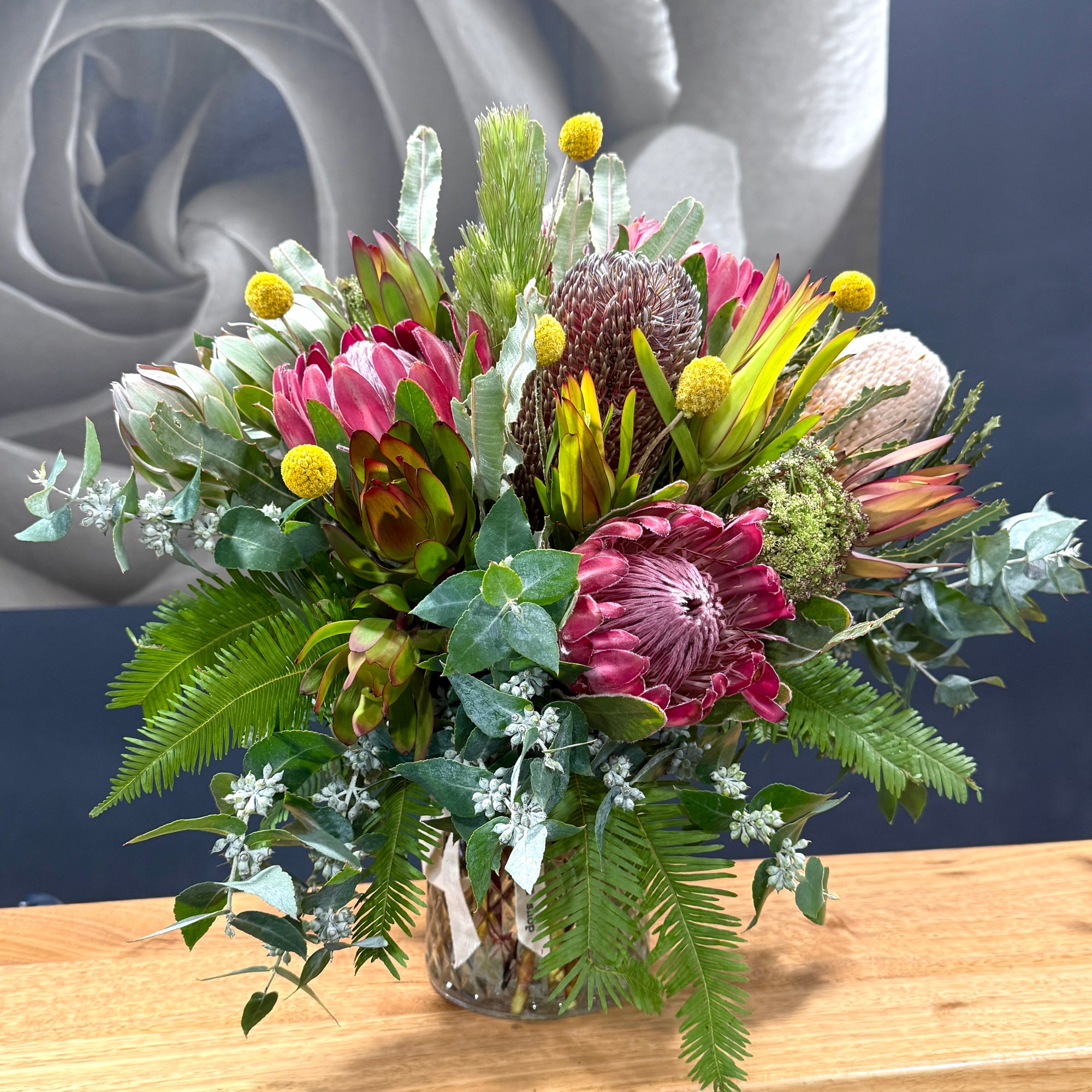 A vibrant mixed floral arrangement in a clear glass vase, featuring prominent pink protea, yellow protea, green ferns, and eucalyptus, displayed on a light wood surface against a dark blue background.