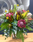 A vibrant mixed floral arrangement in a clear glass vase, featuring prominent pink protea, yellow protea, green ferns, and eucalyptus, displayed on a light wood surface against a dark blue background.