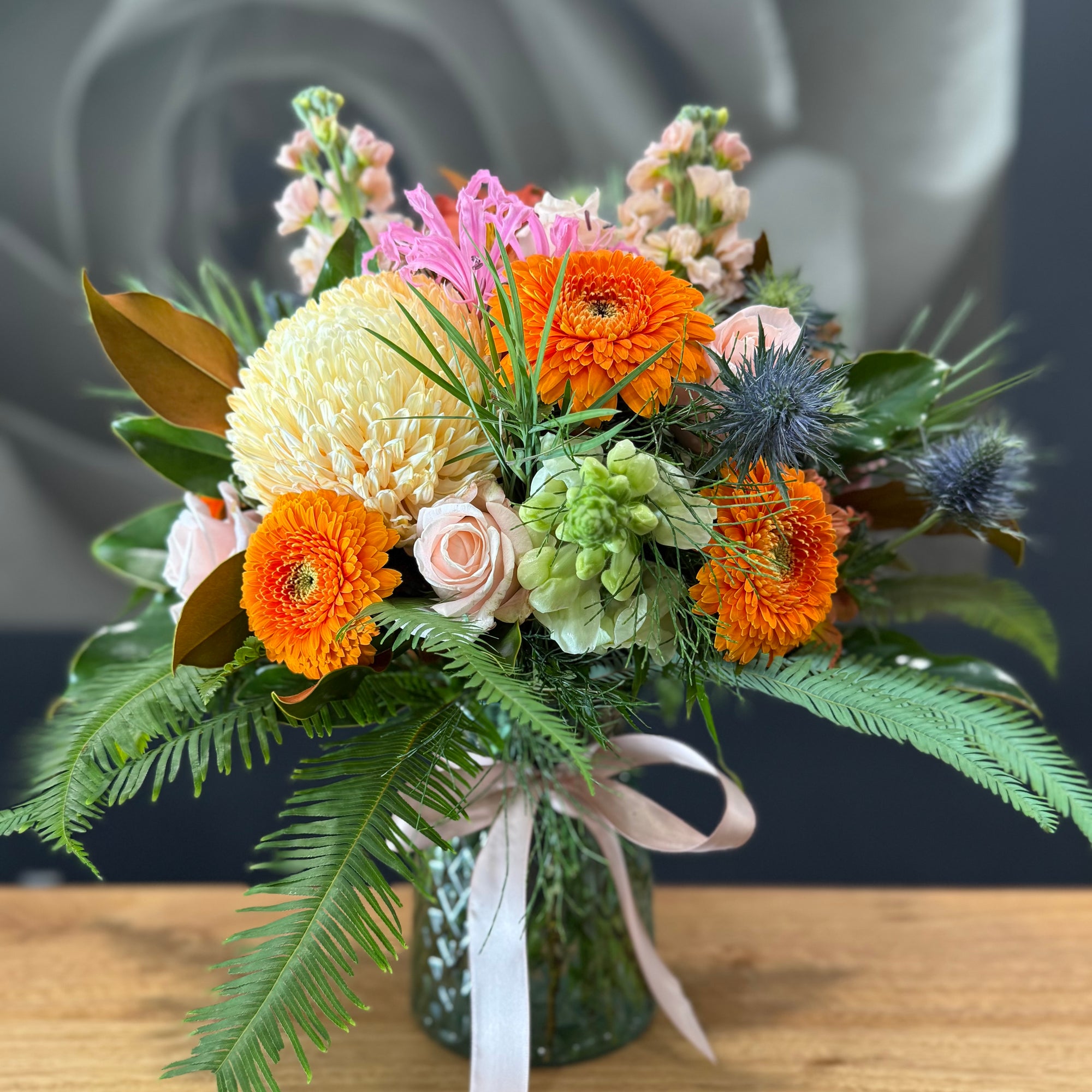 A vibrant  flower arrangement in a textured green glass vase, featuring bright orange gerbera daisies, pale yellow chrysanthemums, pink roses, and green foliage, tied with a white ribbon on a wooden surface.
