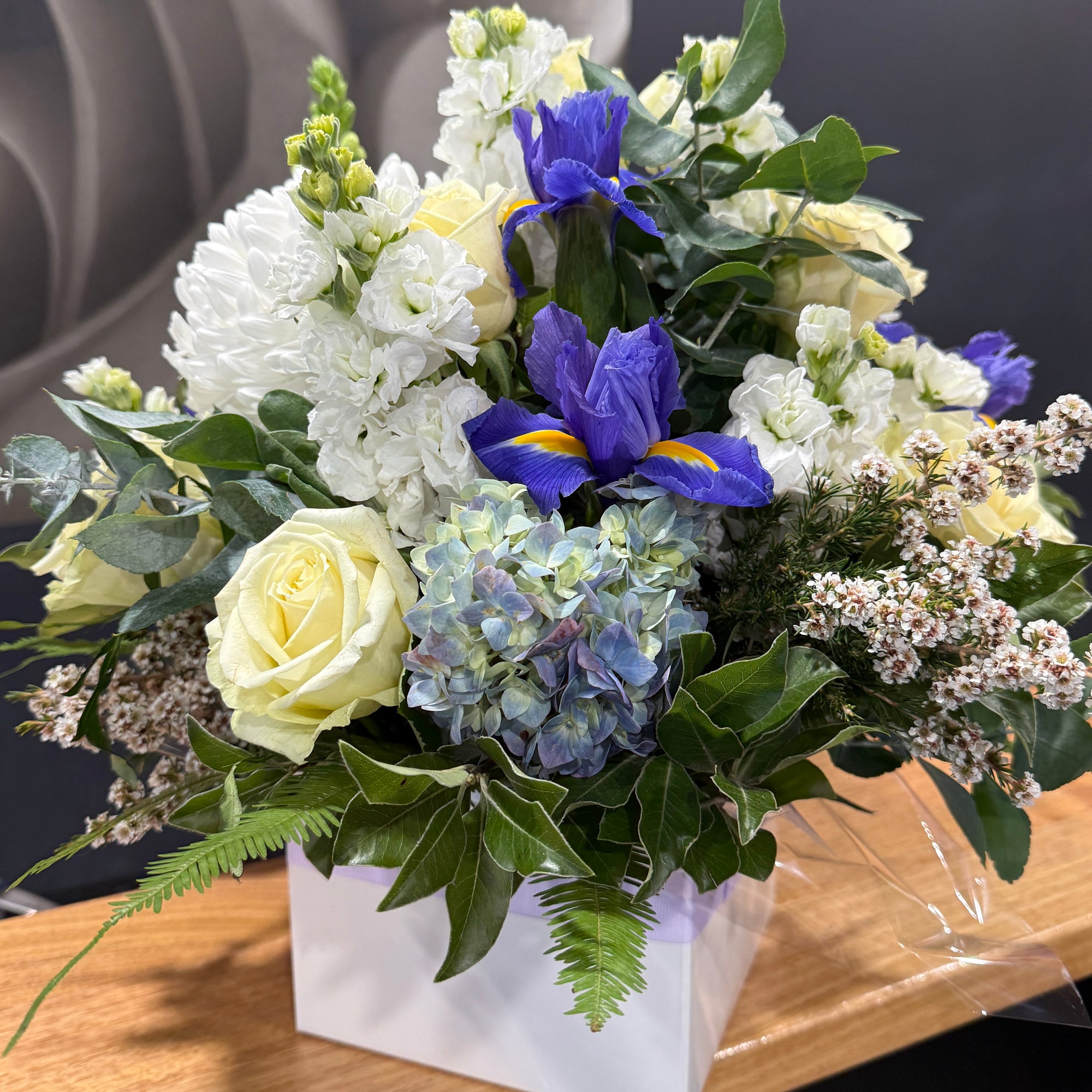 A square floral arrangement in a white box, featuring white and pale yellow roses, blue hydrangeas, purple irises, and green foliage on a wooden surface.